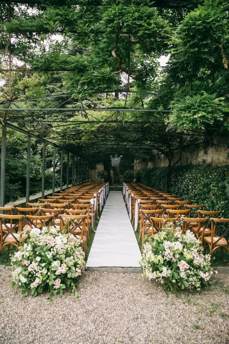 Outdoor ceremony aisle lined with wooden chairs and floral arrangements under greenery at Villa Bossi.