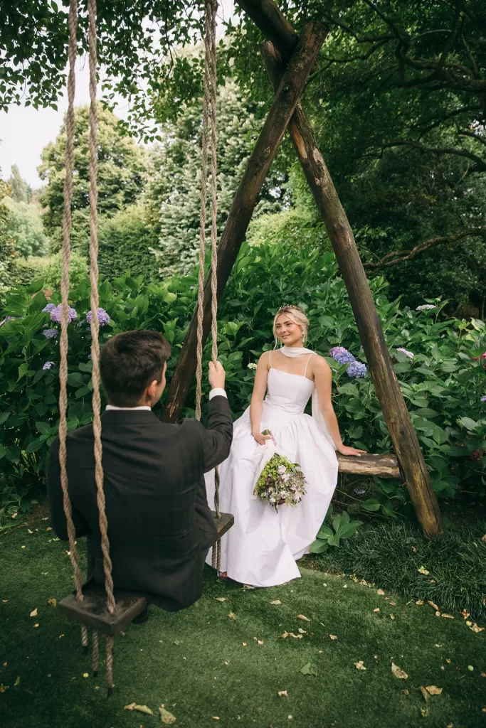 Bride sitting gracefully on a wooden swing while the groom looks on in Villa Bossi’s gardens.