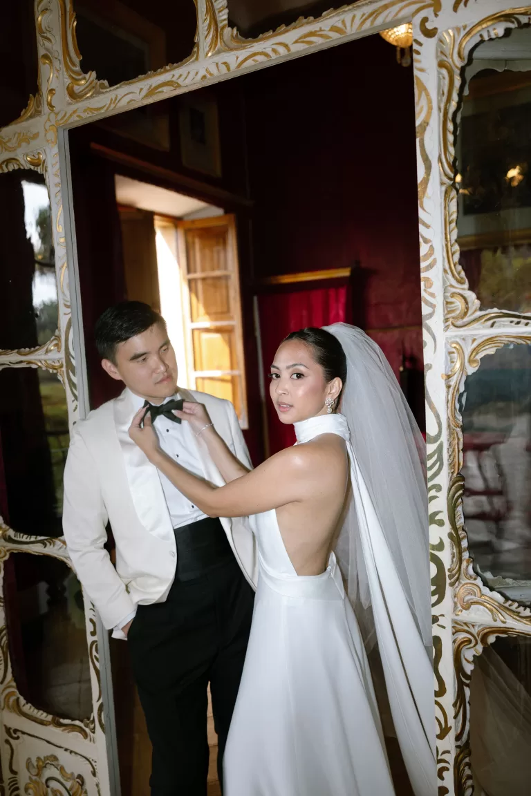 Stylish bride adjusting groom’s bow tie in a vintage room with gilded mirrors, captured by Abraham Garcia.