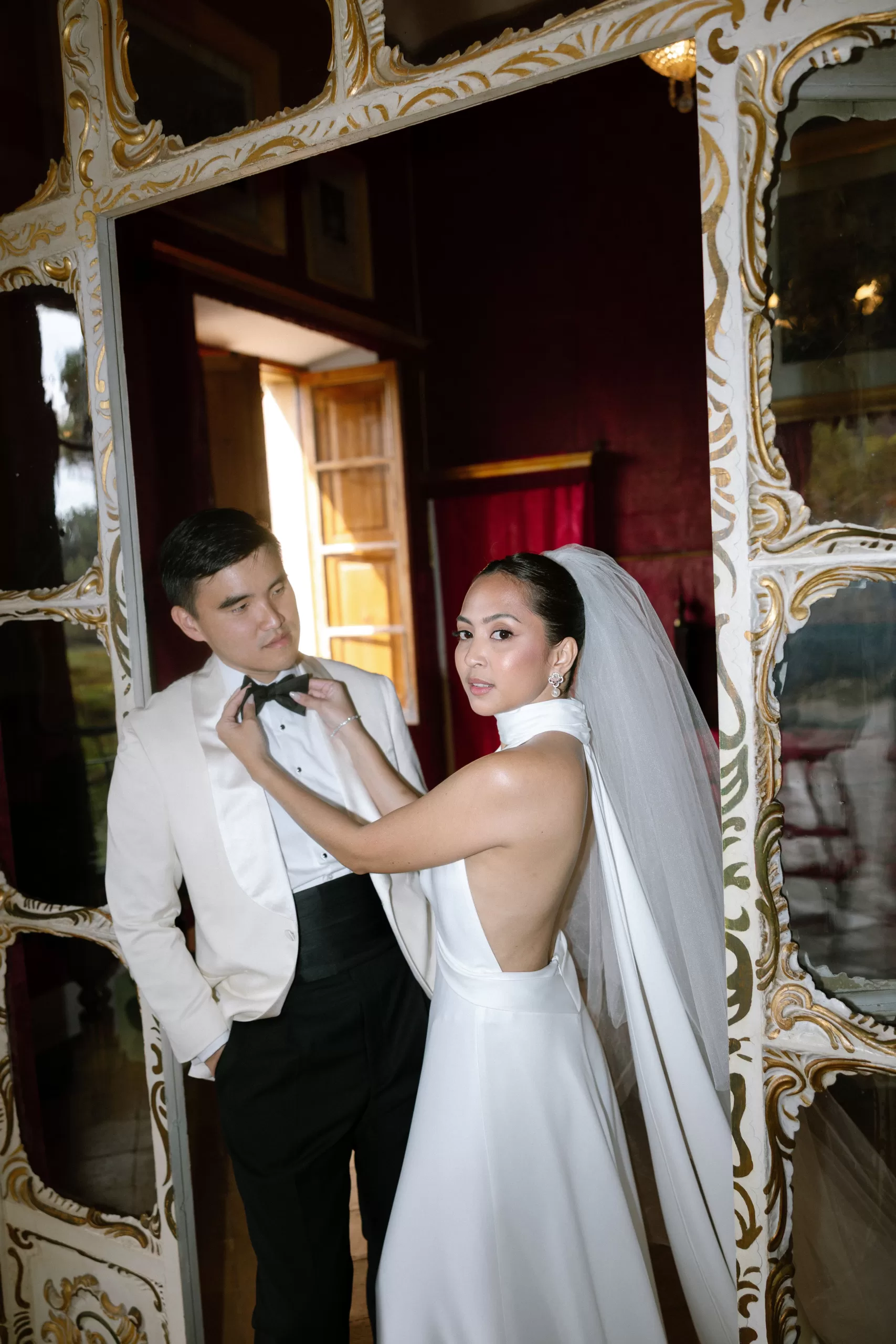 Stylish bride adjusting groom’s bow tie in a vintage room with gilded mirrors, captured by Abraham Garcia.