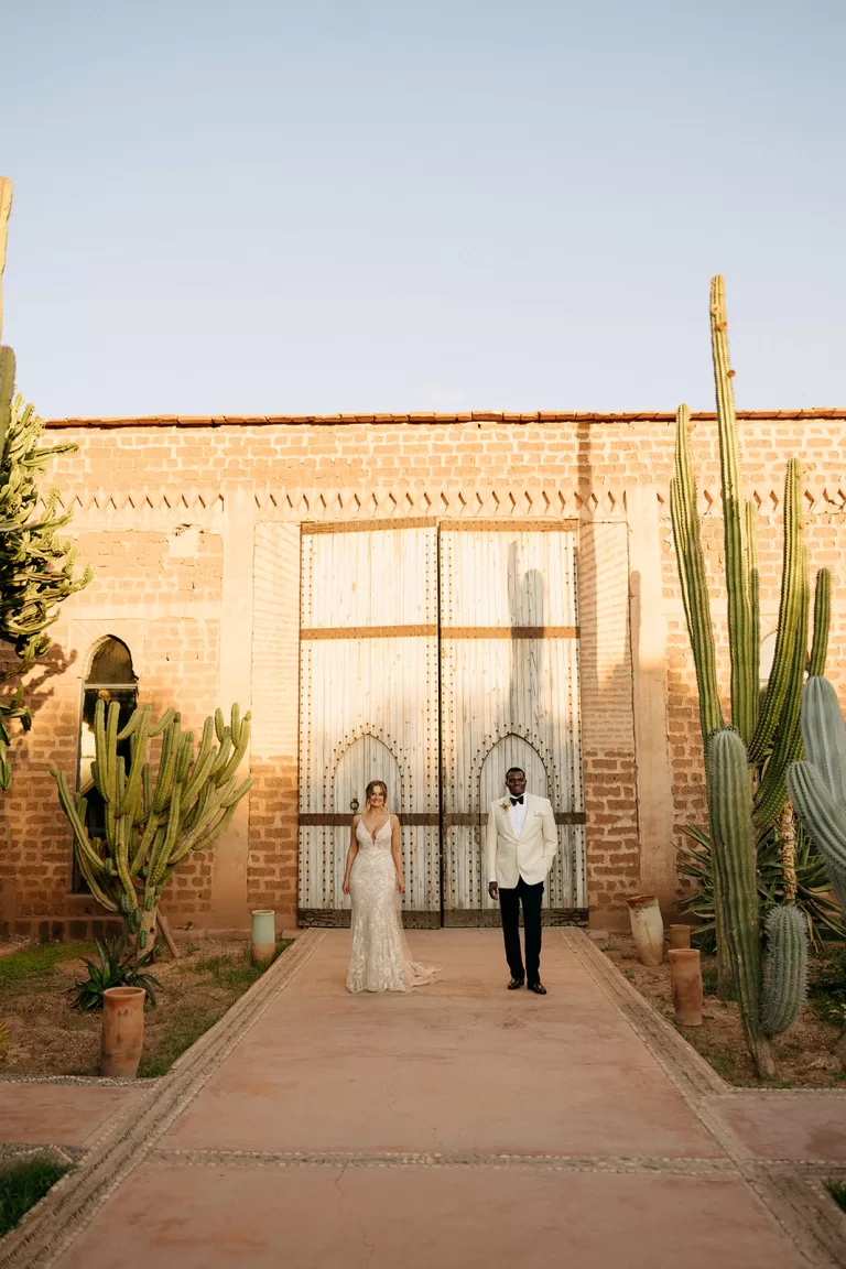 Newlyweds standing together among tall cacti in front of the grand wooden doors at Beldi Country Club.