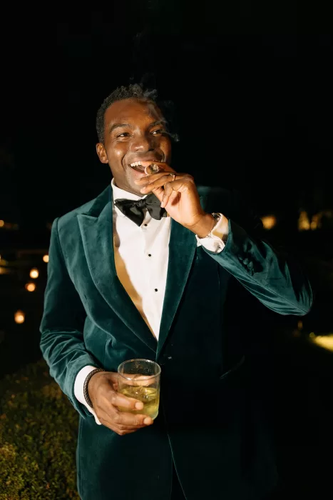 Groom enjoying a cigar and drink at night in a velvet jacket during the Beldi Country Club wedding celebration.