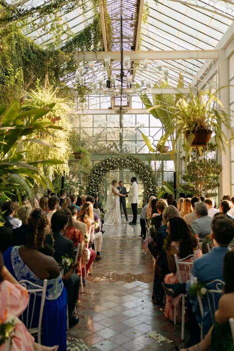 Couple exchanging vows under a floral arch in the glasshouse at Beldi Country Club, surrounded by guests and tropical foliage.