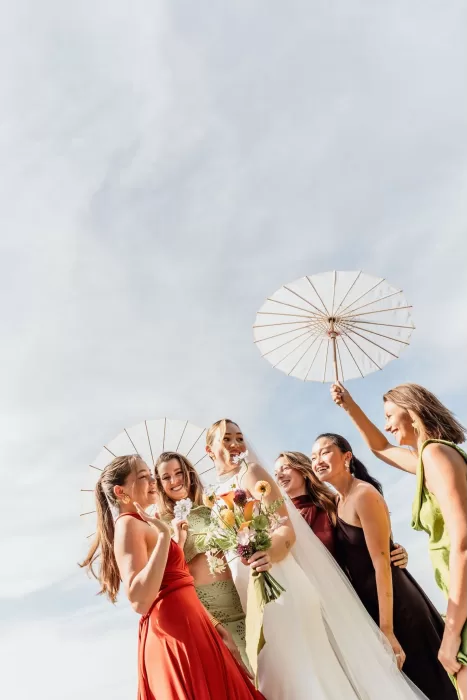 The bride and bridesmaids laughing under white parasols at This Must Be The Place, bathed in bright sunlight.