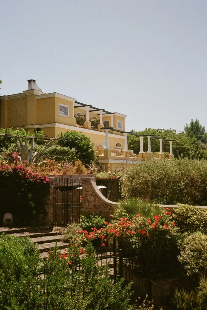 Lush garden terrace leading up to the yellow façade of Caesar Augustus in Capri, surrounded by flowering plants and Mediterranean greenery.