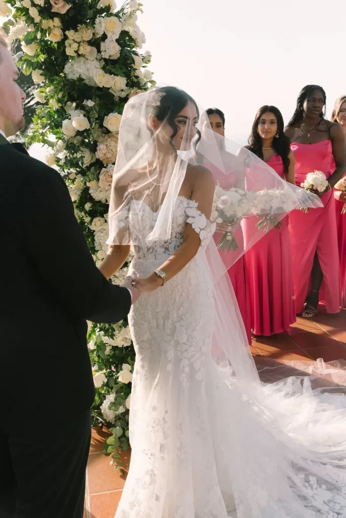 Bride and groom exchanging vows on the terrace of Caesar Augustus in Capri, framed by white roses and pink-clad bridesmaids under the Italian sun.