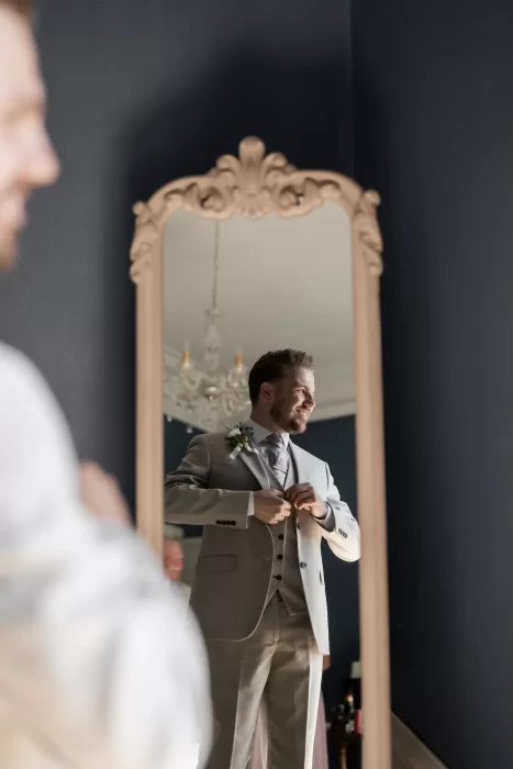 Groom adjusting his waistcoat in front of an ornate mirror while getting ready in a softly lit room at Château de Pordor.