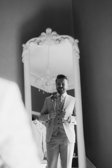 Black and white photo of the groom smiling at his reflection in a vintage mirror before the ceremony at Château de Pordor.