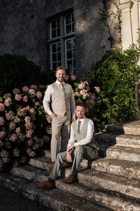 The grooms posing on the château steps beside blooming hydrangeas in the afternoon light at Château de Pordor.