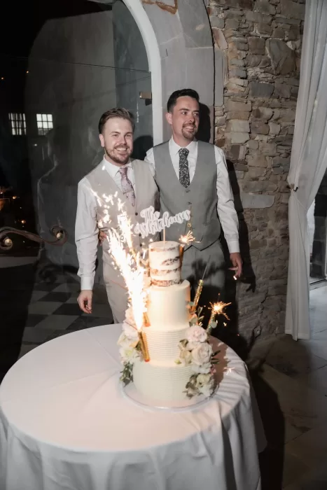 Grooms cutting their sparkler-topped wedding cake inside Château de Pordor’s stone-walled reception room