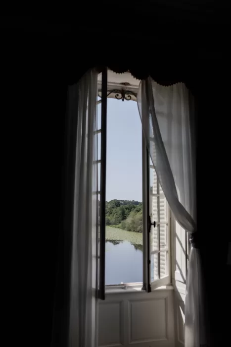 Sunlight filtering through sheer curtains framing a tranquil lake view from a window inside Château de Pordor.