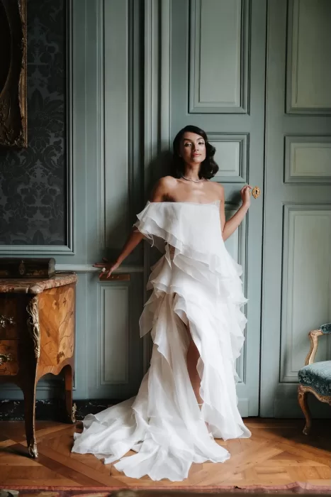Bride posing in a cascading ruffled gown against pale green paneled walls inside Château de Villette.