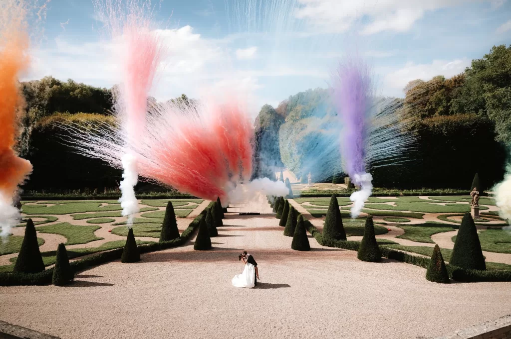 Newlyweds embracing in the gardens of Château de Villette beneath bursts of colorful fireworks in daylight.
