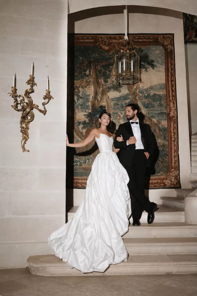 Couple standing elegantly on the staircase at Château de Villette, framed by baroque wall sconces and a French tapestry.