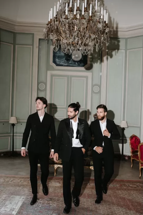 Groom and groomsmen in classic black tuxedos walking confidently under a crystal chandelier inside Château de Villette.