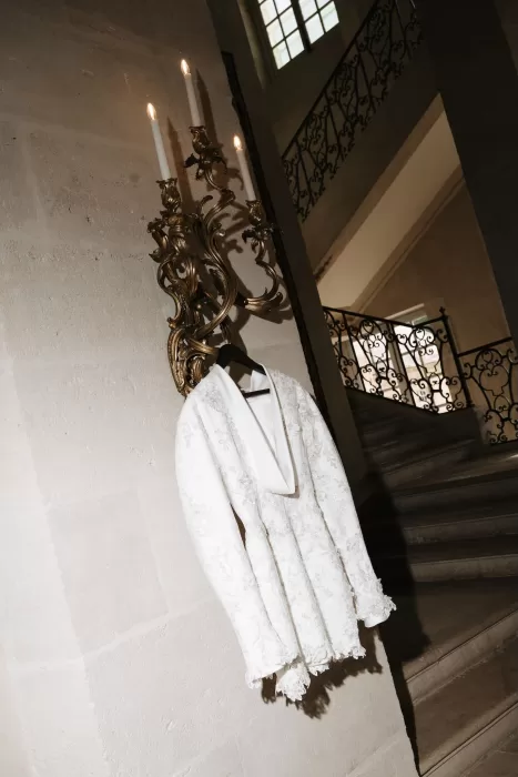 Embroidered white mini wedding dress hanging on a gilded wall sconce inside Château de Villette’s grand staircase.