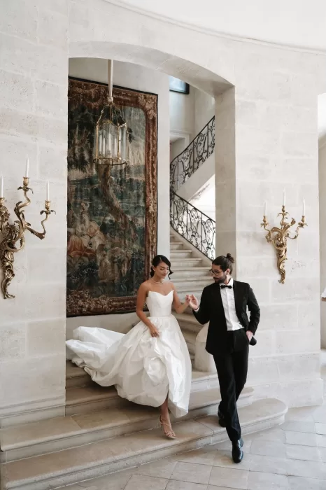 Bride in a structured gown descending the staircase with her groom at Château de Villette, surrounded by ornate gold sconces and tapestries.
