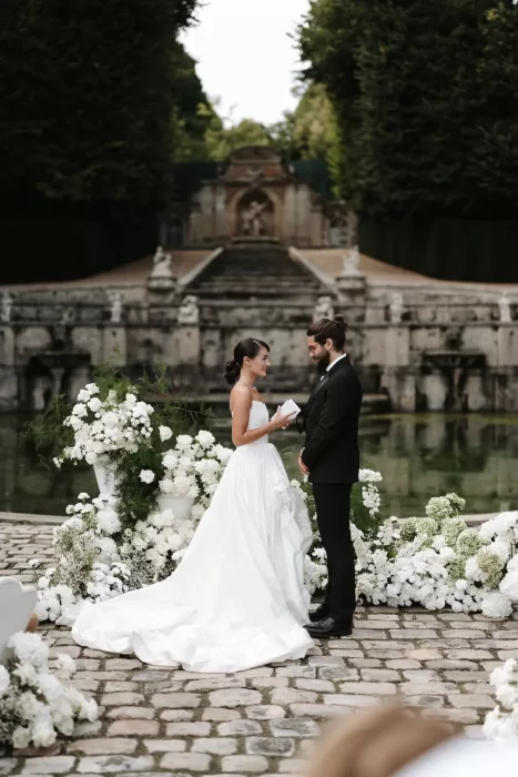 Couple exchanging vows beside the fountain at Château de Villette, surrounded by white floral arrangements.