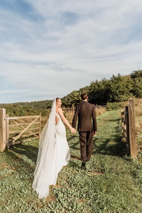 The bride and groom walk hand in hand through a sunlit garden path at This Must Be The Place, surrounded by greenery.