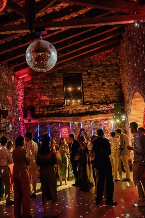 Guests dance beneath a disco ball and colorful lights inside the stone barn at This Must Be The Place.