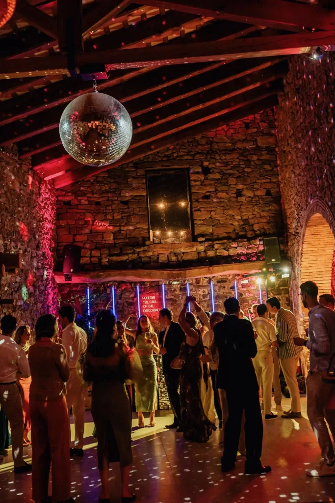 Guests dance beneath a disco ball and colorful lights inside the stone barn at This Must Be The Place.