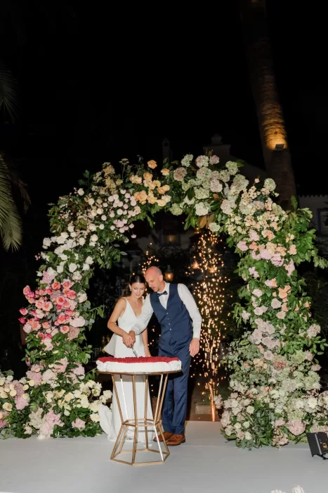Newlyweds cut their wedding cake beneath a floral arch at night, with sparklers glowing behind them at Finca la Concepción.