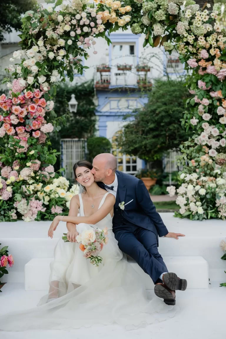 Bride and groom sit smiling beneath a lush floral arch after their ceremony at Finca la Concepción.