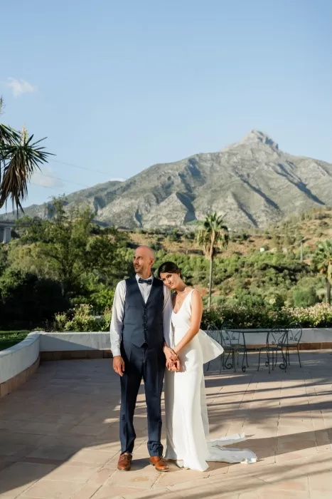 Bride and groom stand arm in arm on the terrace of Finca la Concepción with mountain views behind them.