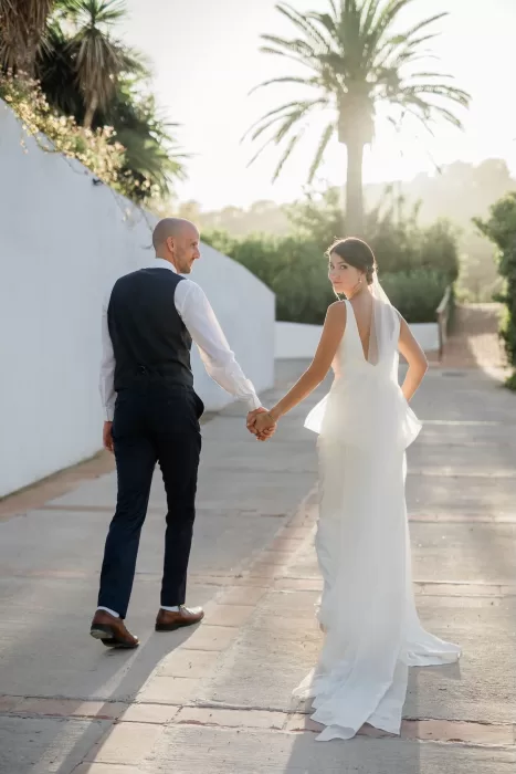 Bride and groom walk hand in hand down a sunlit path lined with palm trees at Finca la Concepción.