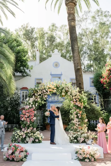 Bride and groom share their first kiss beneath a romantic pastel floral arch in the courtyard of Finca la Concepción.