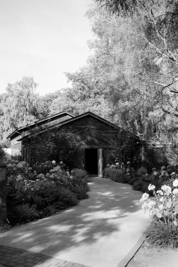 Black and white photo of Hampton Manor’s rustic barn surrounded by lush trees and garden paths.