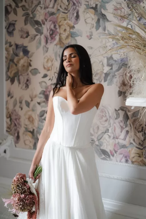 Bride in satin gown standing against a floral wall at Hampton Manor, holding a textured hydrangea bouquet.