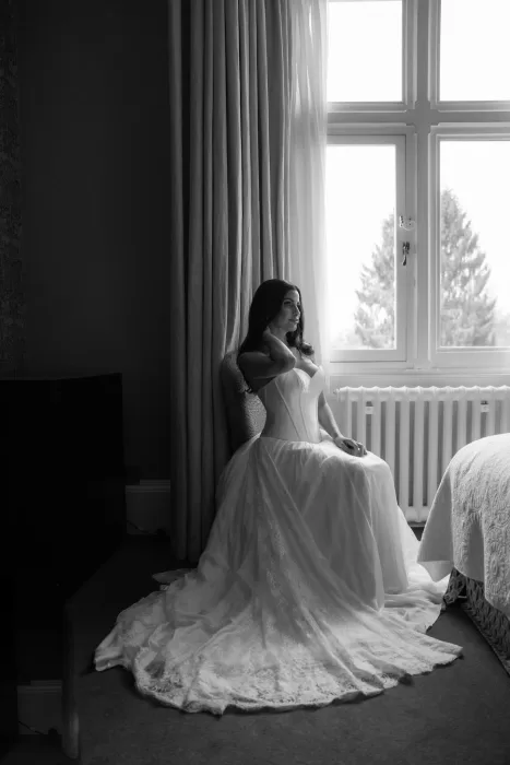 Black and white portrait of the bride sitting by a large window at Hampton Manor, her gown elegantly draped across the floor.