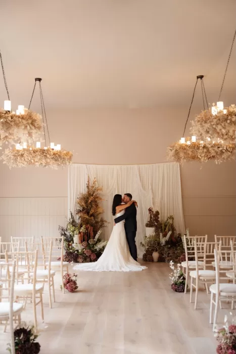 Couple sharing a romantic embrace in Hampton Manor’s ceremony room with floral chandeliers overhead.