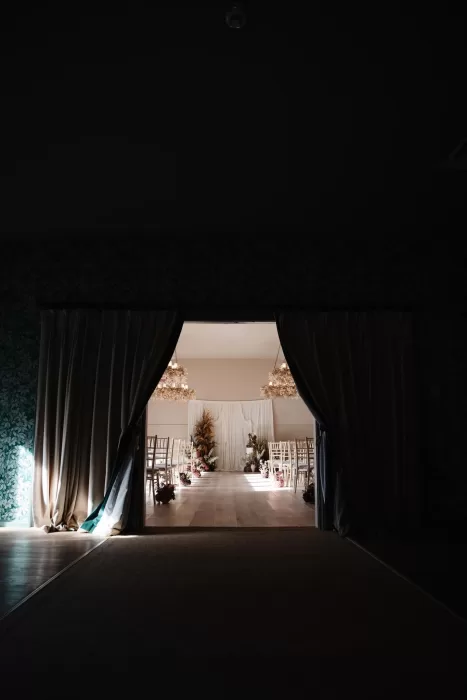 View through dark curtains into the ceremony room at Hampton Manor, softly lit with floral decor and chairs arranged in rows.