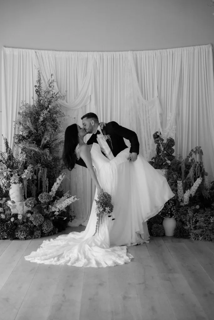 Black and white photo of newlyweds sharing a kiss beneath flowing drapery and floral arrangements at Hampton Manor.