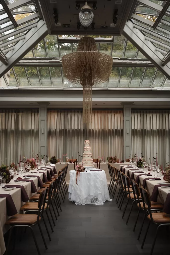 Elegant reception setup at Hampton Manor with long banquet tables, mauve napkins, and a tiered wedding cake beneath a glass ceiling chandelier.
