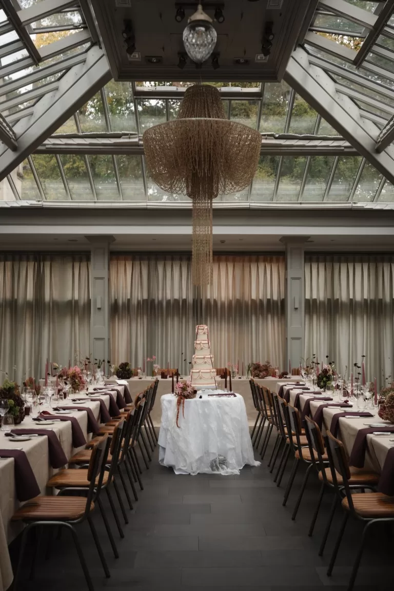 Elegant reception setup at Hampton Manor with long banquet tables, mauve napkins, and a tiered wedding cake beneath a glass ceiling chandelier.