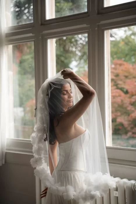 Bride standing by the window at Hampton Manor, softly lifting her floral-edged veil in natural light.