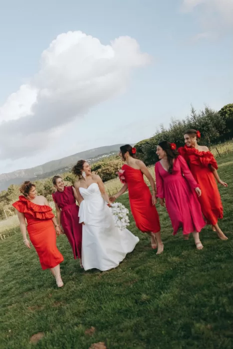Bride and bridesmaids in red and pink dresses laughing together on the lawns of Herdade do Peru.