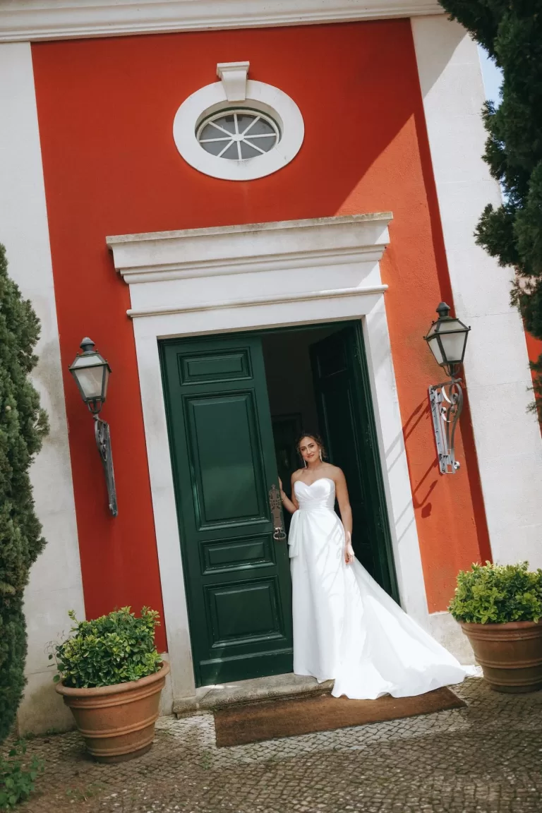 Bride in strapless white gown standing gracefully in the doorway of a red and white villa at Herdade do Peru.