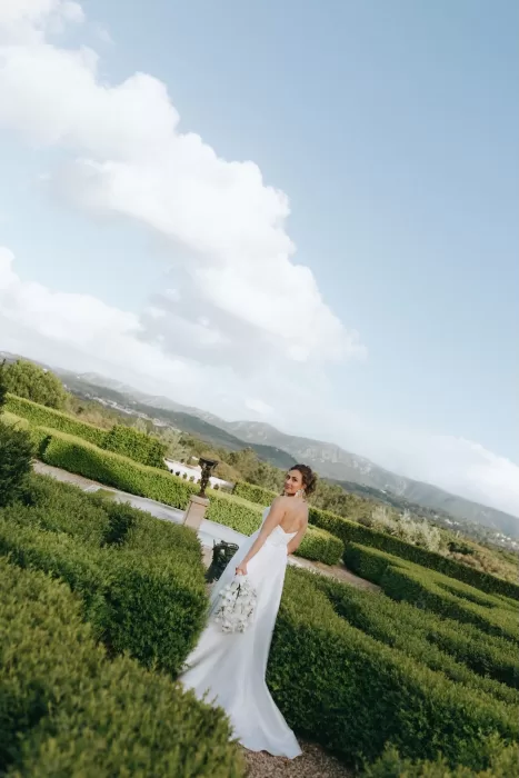 Bride holding white bouquet and looking over her shoulder in manicured gardens with mountain views at Herdade do Peru.