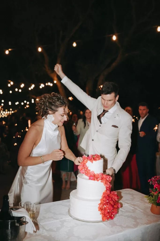 Bride and groom joyfully cutting their wedding cake with red floral decoration during a candlelit reception at Herdade do Peru.