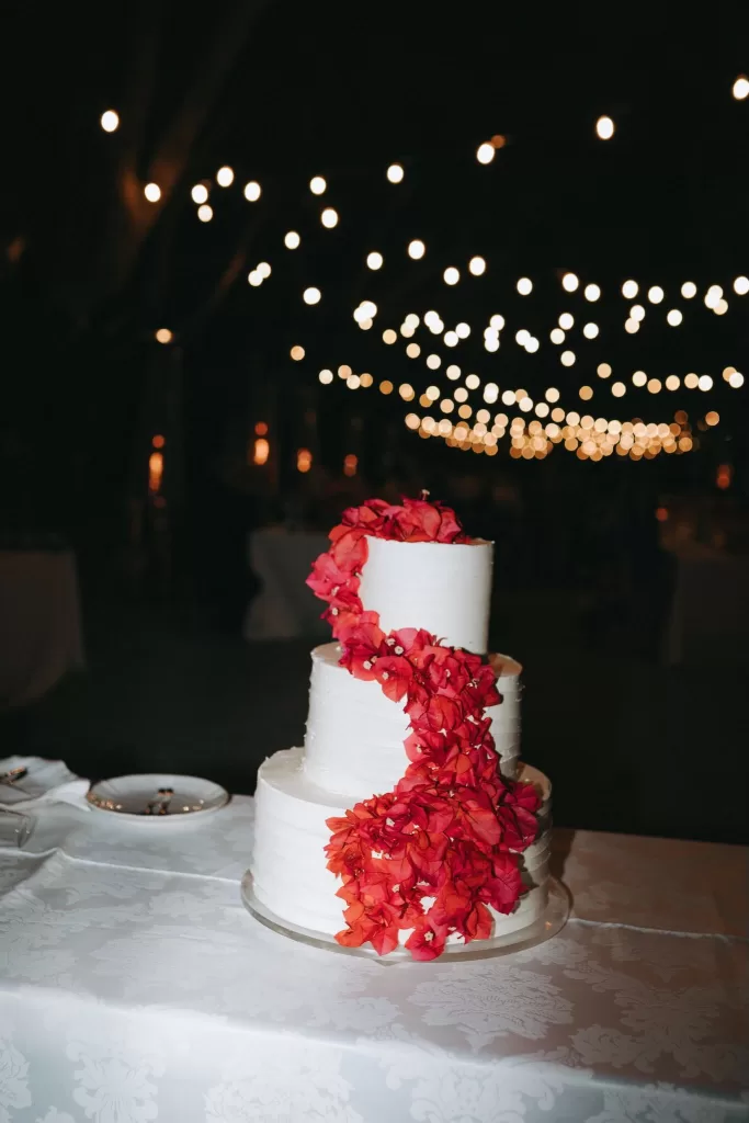 Three-tier white wedding cake adorned with cascading red flowers under twinkling lights at Herdade do Peru.