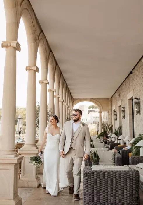 Bride and groom walking hand in hand under the arched colonnade of Hospes Maricel & Spa.
