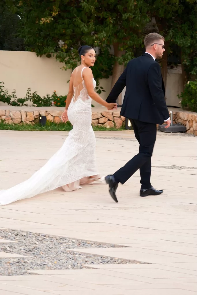 Bride in lace gown walks hand in hand with groom in black tuxedo at Hospes Maricel & Spa.