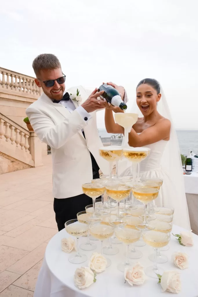 Couple pours champagne into a tower of crystal coupes during their seaside celebration at Hospes Maricel & Spa.