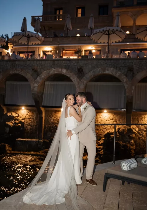 Bride and groom embracing at night by the arches of Hospes Maricel & Spa, softly lit by golden lights.