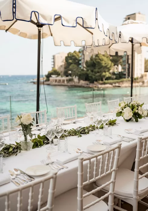 Elegant wedding dining setup overlooking turquoise sea at Hospes Maricel & Spa, with white chairs and floral garlands.