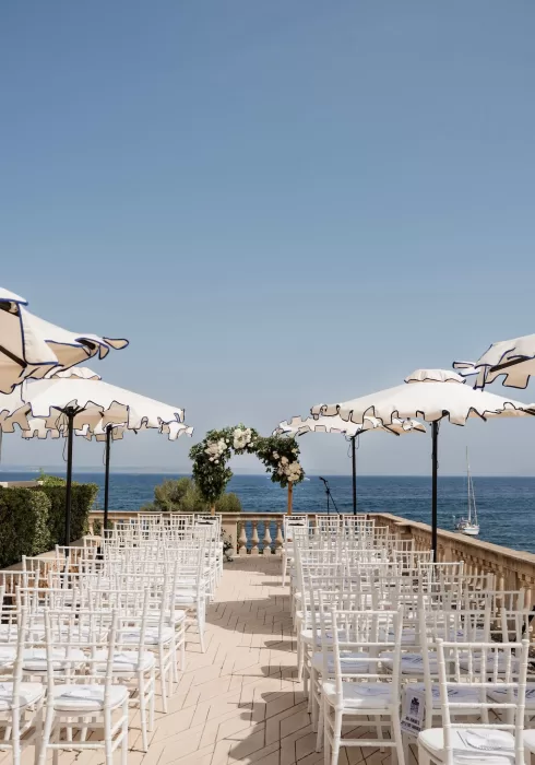 Elegant outdoor ceremony setup at Hospes Maricel & Spa with white chairs, parasols, and a floral arch overlooking the Mediterranean Sea.